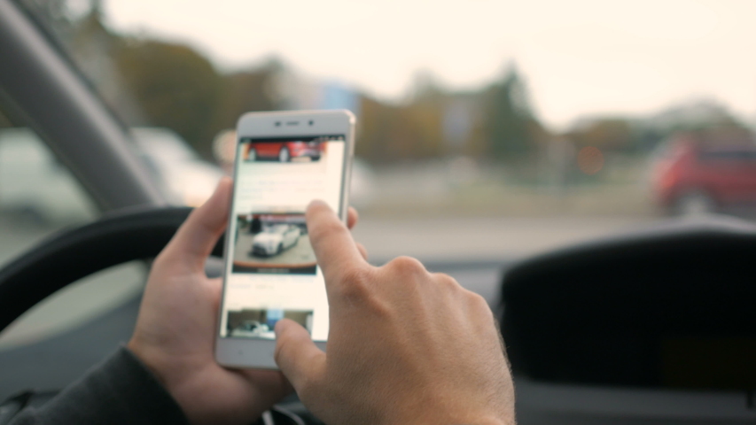 Man chooses to buy a car on the site. Looking at used vehicle to buy on a smartphone app. Driver reads news on mobile phone inside car. Screen is blurred. Focus on finger hand.