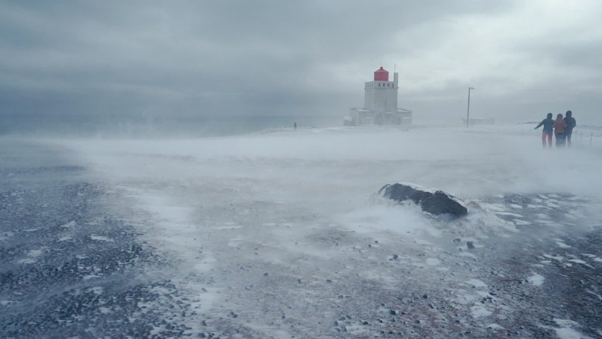 Three people walk towards the Dyrhólaey lighthouse in Iceland on top of the hill during a winter snow storm while it is very windy and the snow blows over the ground.