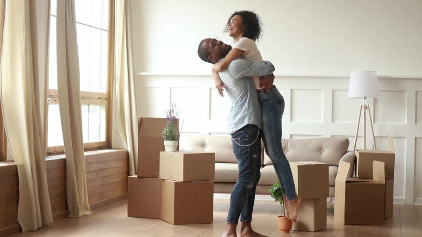 Happy african couple renters owners tenants celebrate moving day in own flat house, excited black man husband holding carrying spinning woman wife having fun among boxes enjoy relocation into new home Royalty-Free Stock Footage #1029780962