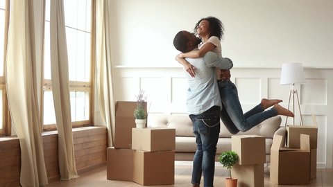 Happy african couple renters owners tenants celebrate moving day in own flat house, excited black man husband holding carrying spinning woman wife having fun among boxes enjoy relocation into new home