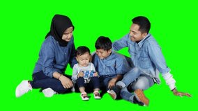 Cute little girl using a mobile phone with her brother and parents while sitting on the floor in the studio. Shot in 4k resolution with green screen background