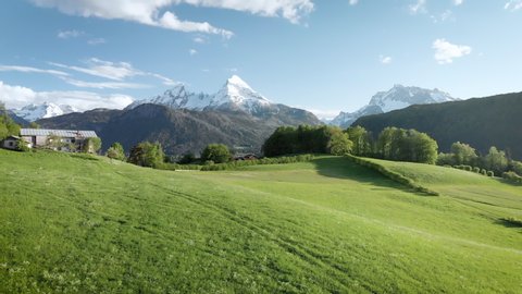 Scenic aerial panoramic view of idyllic rolling hills spring landscape in the Alps with lush green mountain pastures and snow capped alpine mountain tops in the background in evening light at sunset