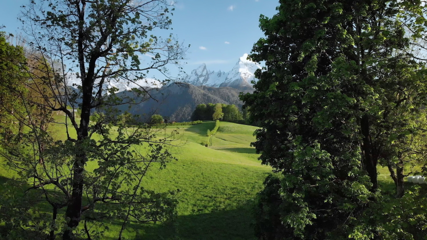 Scenic aerial panoramic view of idyllic rolling hills spring landscape in the Alps with lush green mountain pastures and snow covered alpine mountain peaks in the background in evening light at sunset