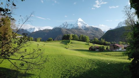 Scenic aerial panoramic view of idyllic rolling hills spring landscape in the Alps with lush green mountain pastures and snow covered alpine mountain peaks in the background in evening light at sunset