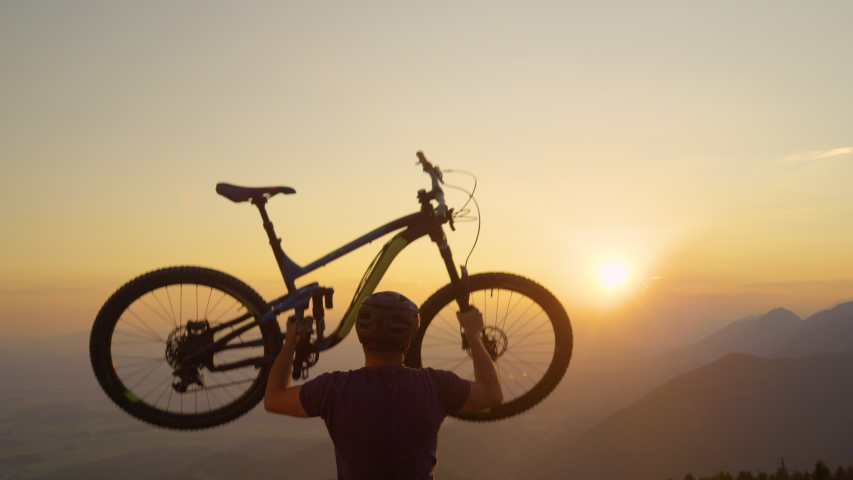 SLOW MOTION, SUN FLARE, CLOSE UP: Happy man lifts his bicycle above his head at sunset after a mountain biking trip in the beautiful mountains. Cheerful tourist celebrates winning a mountain bike ride Royalty-Free Stock Footage #1029829778
