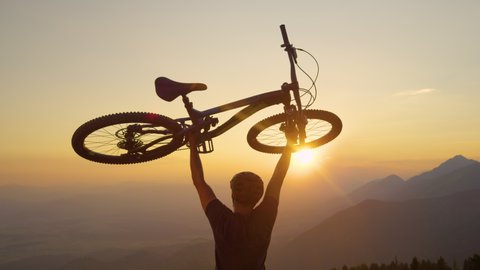 SLOW MOTION, SUN FLARE, CLOSE UP: Happy man lifts his bicycle above his head at sunset after a mountain biking trip in the beautiful mountains. Cheerful tourist celebrates winning a mountain bike ride