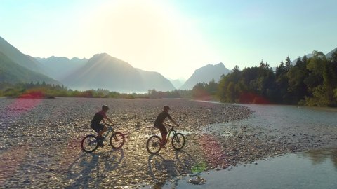 AERIAL SUN FLARE: Beautiful sunlit landscape surrounds two male travelers riding electric bikes along the calm river. Flying along men riding electric bicycles around the spectacular Soca river valley