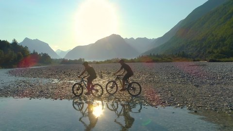 DRONE, LENS FLARE, SILHOUETTE: Golden sun rays shine on two travelers exploring the picturesque Soca river valley on ebikes. Spectacular nature surrounds guys riding electric bikes along calm river.