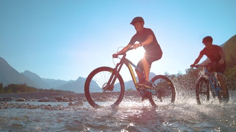 SUPER SLOW MOTION, LOW ANGLE: Guys riding ebikes along the shallow river and splashing refreshing water on a sunny day in beautiful Slovenian nature. Young friends spraying water during a bike ride.