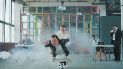 Office chair race. Young businessman is riding a chair in the office like a winner. His colleague helps him. Smoke screen. Office life. Business team. Office party