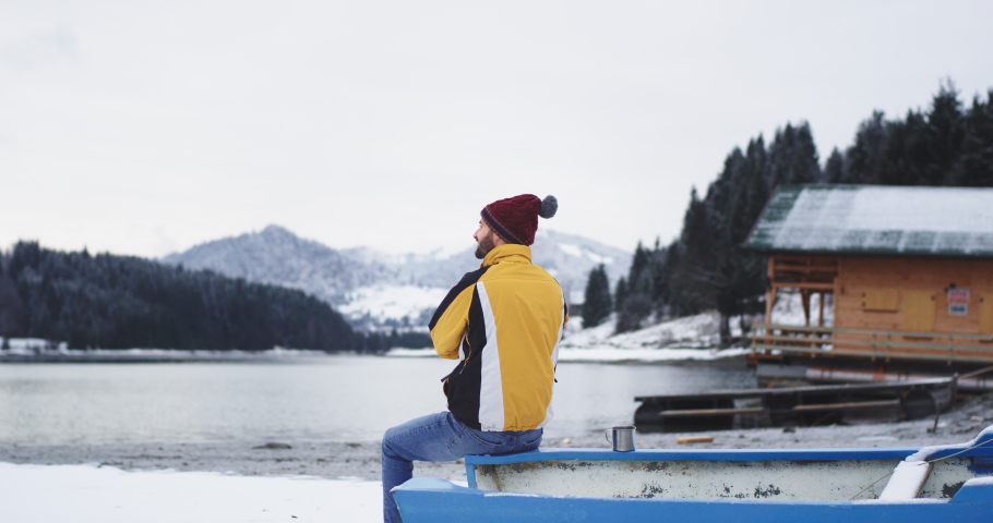 Beside of a big snowy lake and forest charismatic young tourist admire the view he sitting beside of a blue boat and turning to the camera and smiling large , tourist wearing a orange jacket and red