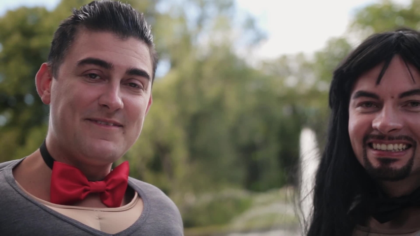 Bearded funny guy with long dark hair and young man with short hair are dressed in fake body muscle costume and bow ties, they are dancing and singing on camera standing outside at some park.