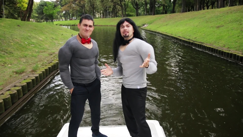 Two handsome white dark-haired funny guys wearing fake muscle chest and arms costumes are energetically dancing on camera standing in boat which moves along narrow river at some green park.
