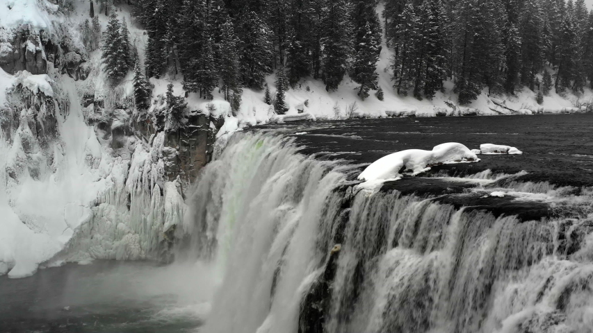 Aerial flyover view of river and waterfall in winter near trees / Island Park, Idaho, United States