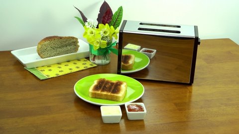 A man at the breakfast table is spreading butter and jam on a browned toast made from the white bread, then puts the toast on the green plate. 