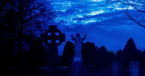 Hyper lapse of clouds moving over jesus statue and celtic cross in dark spooky cemetery at night