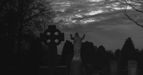 Hyper lapse of clouds moving over jesus statue and celtic cross in dark spooky cemetery at night monochrome