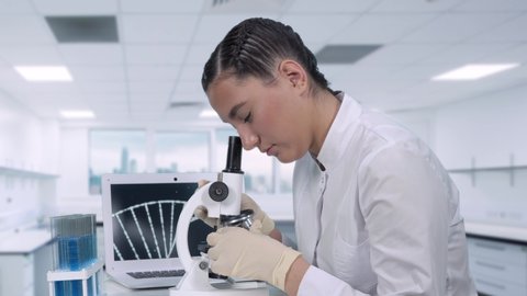 A female lab technician sitting at a table next to a laptop in a modern medical laboratory looks at biological samples under a microscope. A sample of DNA is being examined. 库存视频