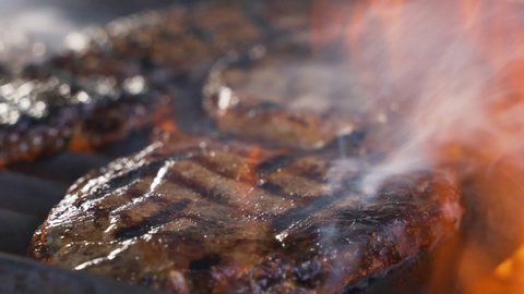Chief using the tong and turning the meat on the grill. Fire is burning, hot juicy oil steak cooking close up slow motion. Grill, tasty beefsteak slow motion close up