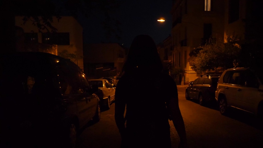 Night shot of a woman walking down a road with parked cars on both sides.