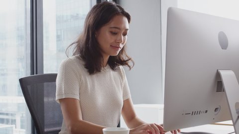 Millennial Asian woman sitting at an office desk using a computer and drinking coffee, waist up