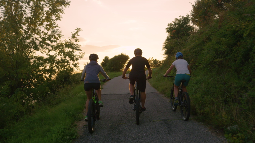 SLOW MOTION, LENS FLARE: Unrecognizable cyclists pedal along the empty road at golden sunset. Three young friends enjoy a scenic bicycle ride in the Slovenian countryside on a sunny summer morning. Royalty-Free Stock Footage #1029925598