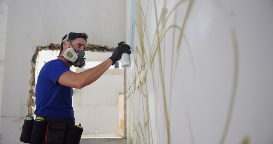 Side view of Caucasian graffiti artist painting with aerosol spray on the wall. He is wearing protective mask
