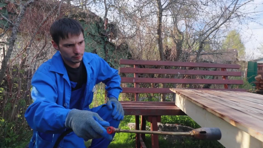 A man with a gas torch burns a table of boards and grinds it with emery sandpaper. Prepares under the cover of yacht varnish. Work outdoors.