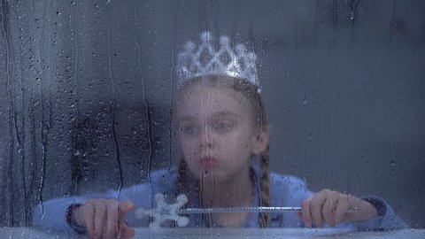 Upset little girl in crown with magic stick sitting alone behind rainy window