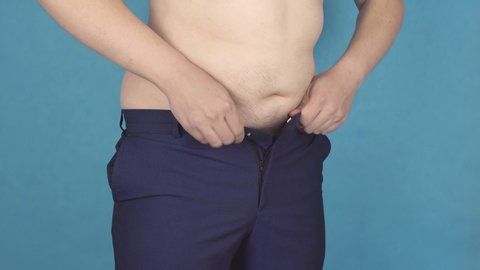 Fat young guy with a big belly fastens tight pants in his waist concept of overweight and obesity, blue background, abdomen