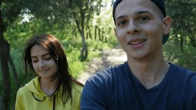 Handsome man with beautiful young girl walking on the road in the park and doing video selfie among green grass and row of trees. Joyful couple shooting video blog. Slow motion