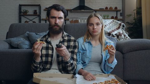 Young couple sitting on the floor near the sofa, holding pizza and watching TV. No emotions, communication problem.