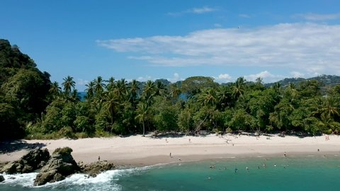 Aerial view of turquoise sea water and people walking in tropical rocky beach with jungle vegetation, waves reaching the sand in Manuel Antonio national park, Costa Rica. Pull out shot.