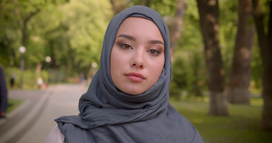 Portrait of muslim woman in hijab with bright make up watching intently into camera walking in the park.