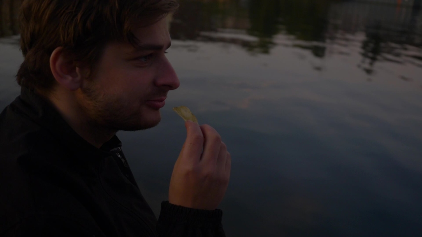 Video of a man nodding eating potato chips contently on the edge of a pier over a relfective lake during the evening
