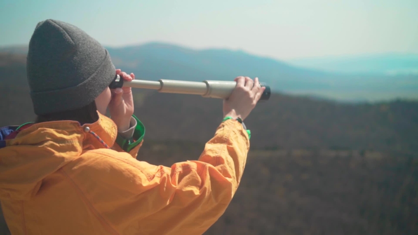A young girl with long dark hair in a yellow jacket and a gray cap is standing in the mountains and looking through a telescope. Background mountains, sky.