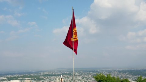 Flag of Kyrgyzstan waving in the wind with some clouds and blue sky. In the background the city of Osh can be seen.