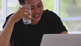 Slow motion scene video of Asian man sitting, smiling, drinking a glass of milk and using laptop in living room.

