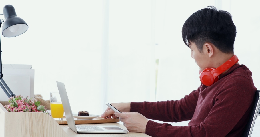 Asian man wearing red headphone, eating chocolate cake during using cellphone, there is laptop on table in living room.
