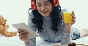 Close up scene video of beautiful Asian woman wearing red headphone during using tablet and drinking orange juice in bedroom.

