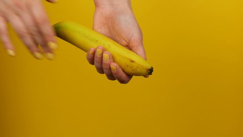 Female hand with yellow manicure holds a ripe banana fruit on a yellow background