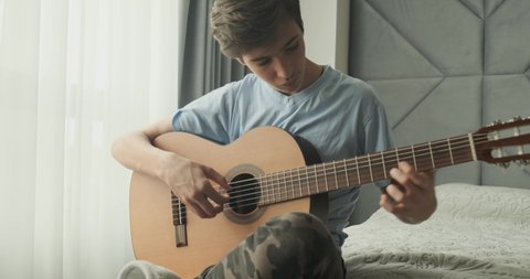 Young man playing on acoustic guitar, at home. Teenage boy with a wooden guitar sits on a sofa.  Child playing guitar.  Teen guy plays on a classical guitar. Real time footage. 