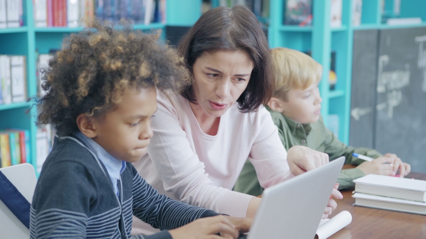 Tracking right shot of middle aged female teacher and group of diverse elementary students sitting at desk in classroom. Tutor explaining mixed race schoolboy how to work on laptop computer Royalty-Free Stock Footage #1030053032