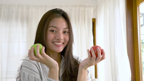 portrait of a young lovely pretty asian girl biting an green apple in white kitchen in morning. Her hands holding green and red apple that made her strong and healthy