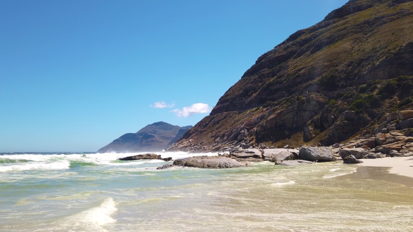 Ocean waves come in at Noordhoek beach in South Africa. Green mountain and blue sky in the background. Slow pan.