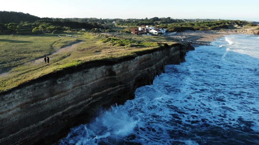 Aerial descending 4k drone shot of two guys standing on the edge of a cliff, and surfers walk by, while waves are breaking in, on a sunny afternoon Mar del Plata, Argentina.