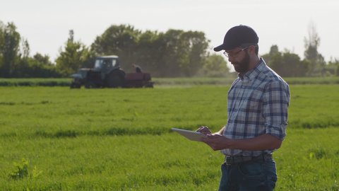 A young bearded farmer in a cap works on the field in spring, use a digital tablet. In the background, the tractor moves out of focus. Farmer using digital tablet. Concept of technologies.