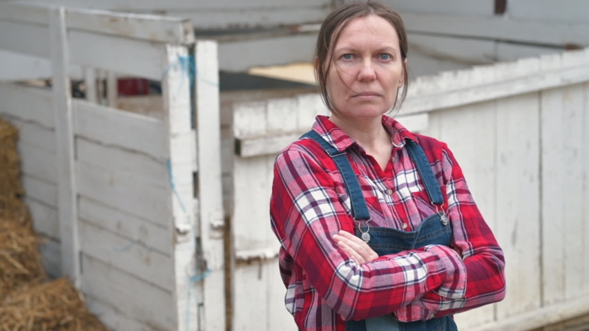 Serious female farmer posing on farm. Confident woman farm worker wearing plaid shirt and jeans overalls looking at camera, arms crossed