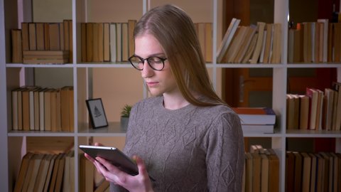 Closeup portrait of young attractive female student in glasses using the tablet and showing green chroma screen to camera in the college library indoors