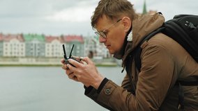 Mature Man wearing glasses in the city on the embankment of the river controls the drone using a remote control, making aerial shooting. Close up portrait. Cloudy weather, spring or cold summer